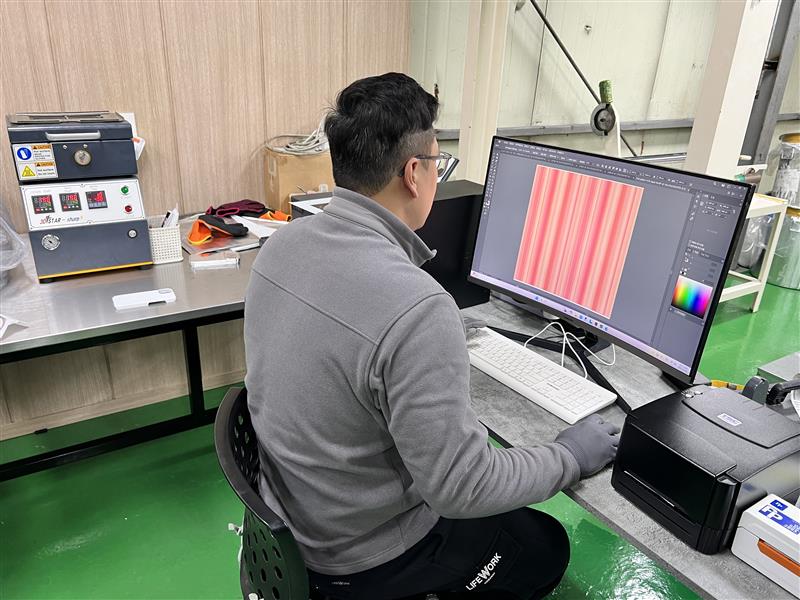 Man working at a curved monitor editing a pink and red striped textile pattern in design software, seated at a workstation in a factory setting with lab equipment on the table beside him.