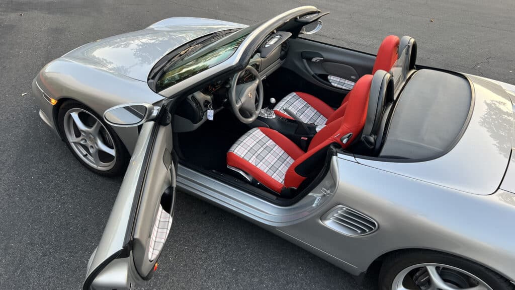Silver Porsche Boxster convertible with driver and passenger doors open, showing red leather seats with white and red plaid inserts and matching door panels.