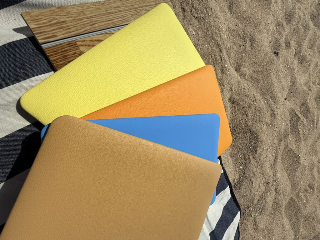 Fan of tan, blue, orange, and yellow leather panels laid on a striped beach towel and wood plank, with sand visible in the background.