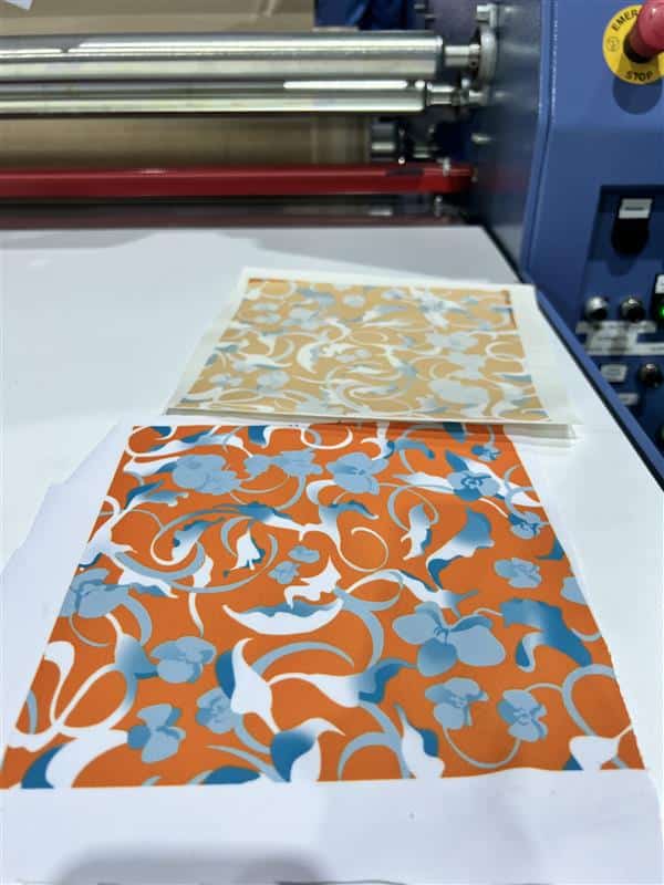 Two floral print fabric swatches, one in orange and blue and one in tan and blue, resting on a printing machine's work surface next to its rollers and control panel.