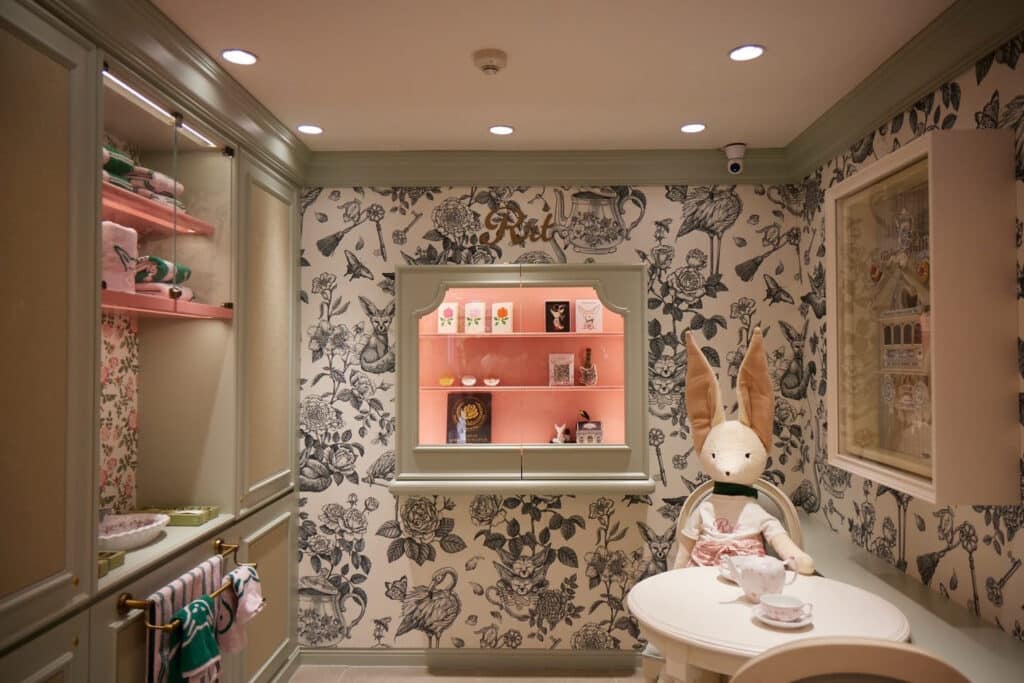Whimsical retail display room with black and white floral toile wallpaper, sage green built in cabinetry, a pink lit display case with figurines, and a plush rabbit character seated at a small tea table.