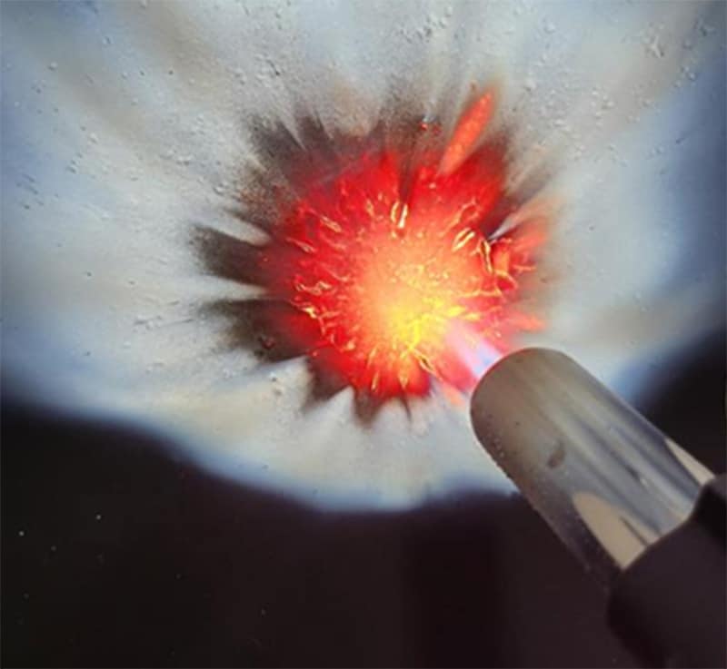 Close up of a flame retardancy test on white upholstery fabric, showing a torch applying direct flame with a scorched red and orange burn zone and light charring around the edges.