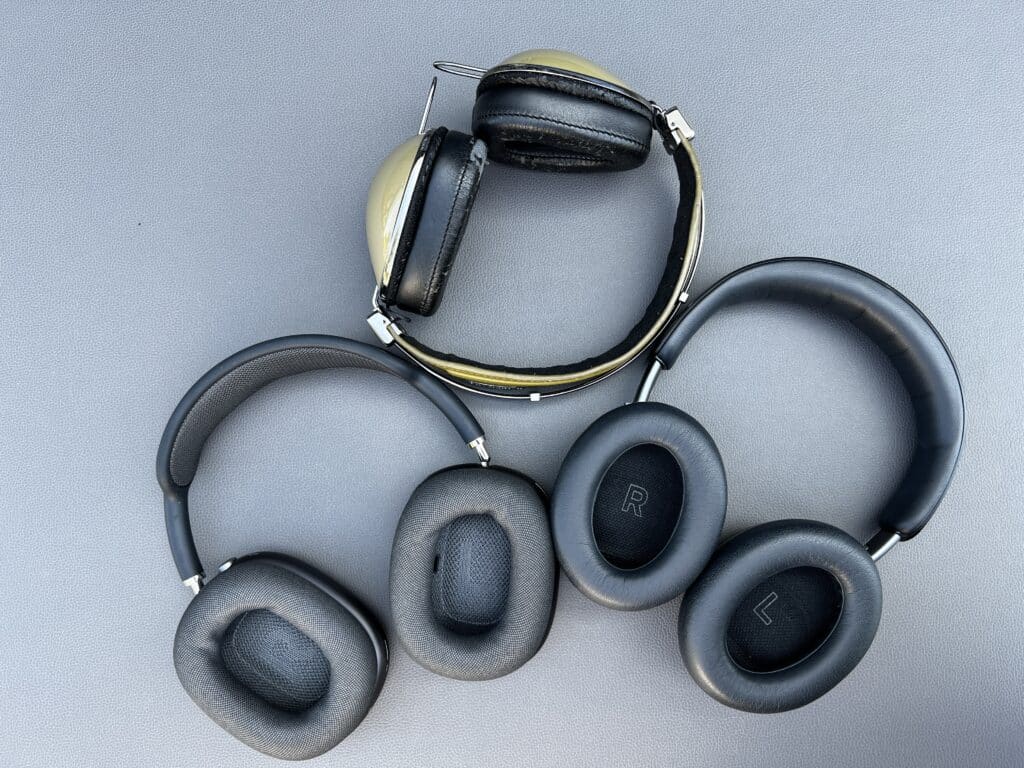 Three pairs of headphones with leather ear cushions and headbands laid on a gray leather surface, including a gold and black vintage style pair and two modern noise cancelling pairs marked L and R.