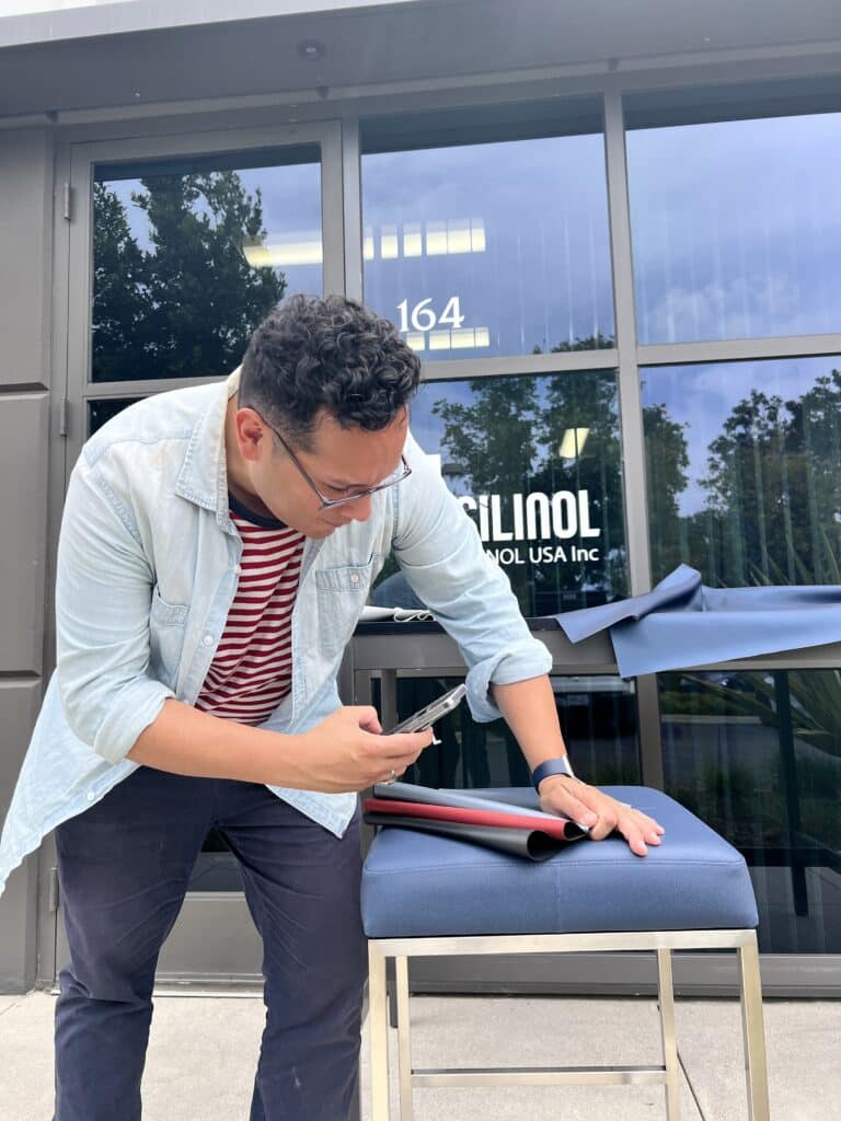 Man in a denim shirt photographing rolled red, black, and gray leather swatches on a blue stool outside the Silinol USA storefront at 164.