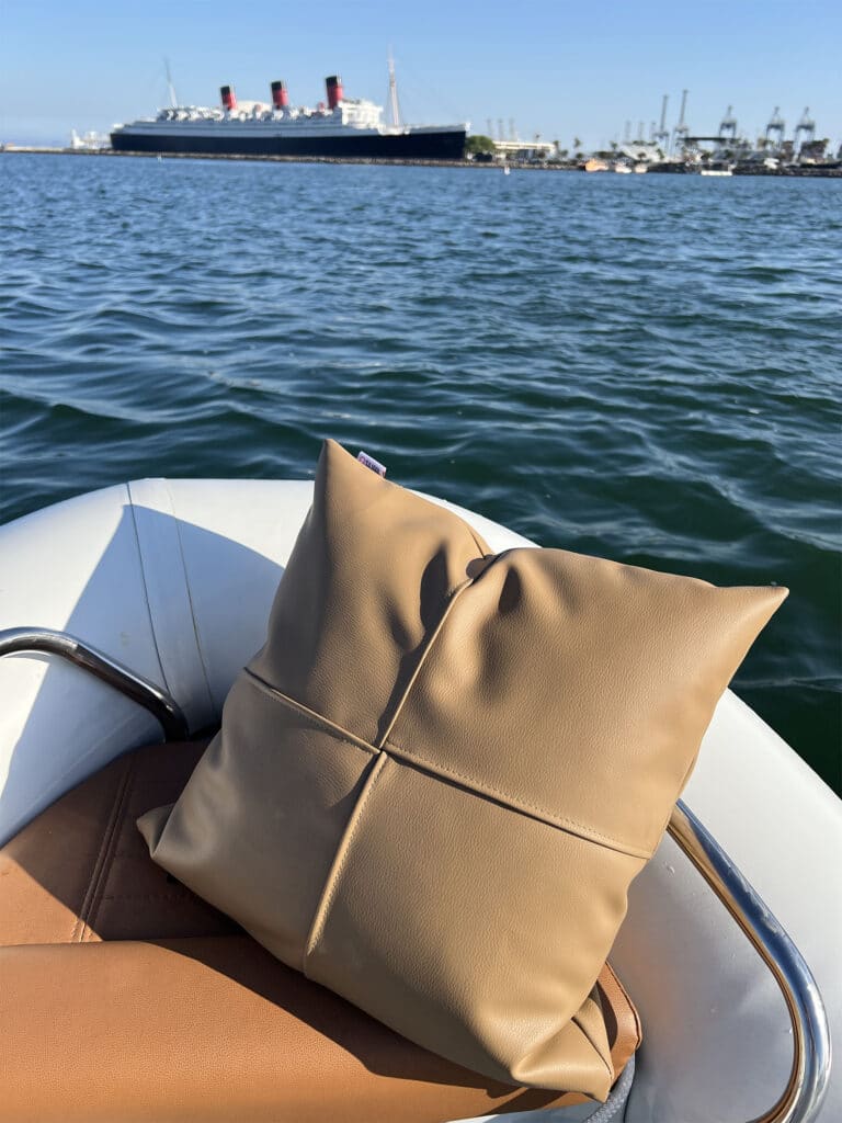 Tan leather cross stitched pillow with a Silinol tag resting on a boat's tan seat cushion, with the Queen Mary ocean liner visible across the harbor in the background.