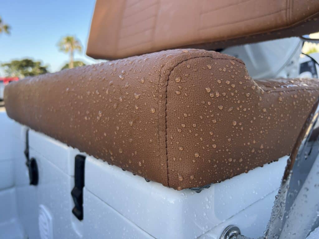 Close up of water beaded up on brown leather boat seating, showing water resistance on the bench edge and backrest against a blurred marina and palm tree background.