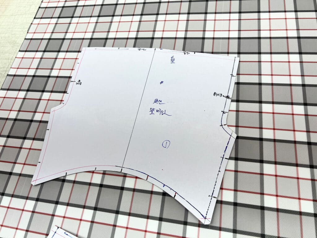 Paper cutting pattern with Korean handwritten notes and pink and blue guide lines laid on top of gray, red, and white plaid fabric ready for cutting.