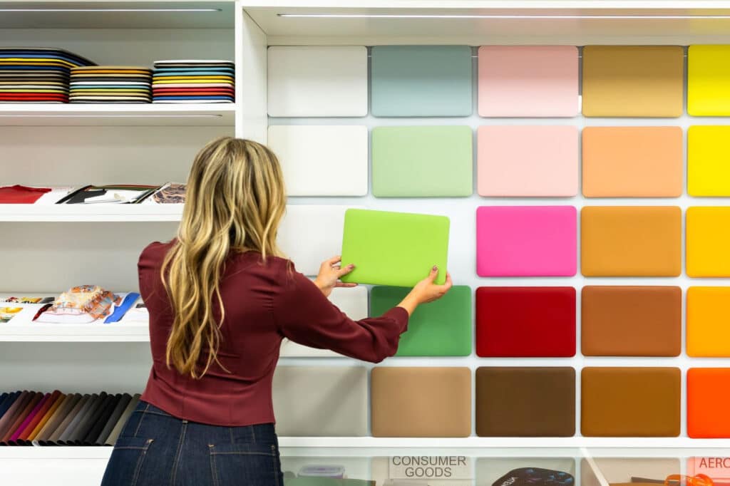 Woman selecting a green color swatch from a large wall display of leather color panels in a studio, with rolled fabric samples and folded product on shelves to the left.