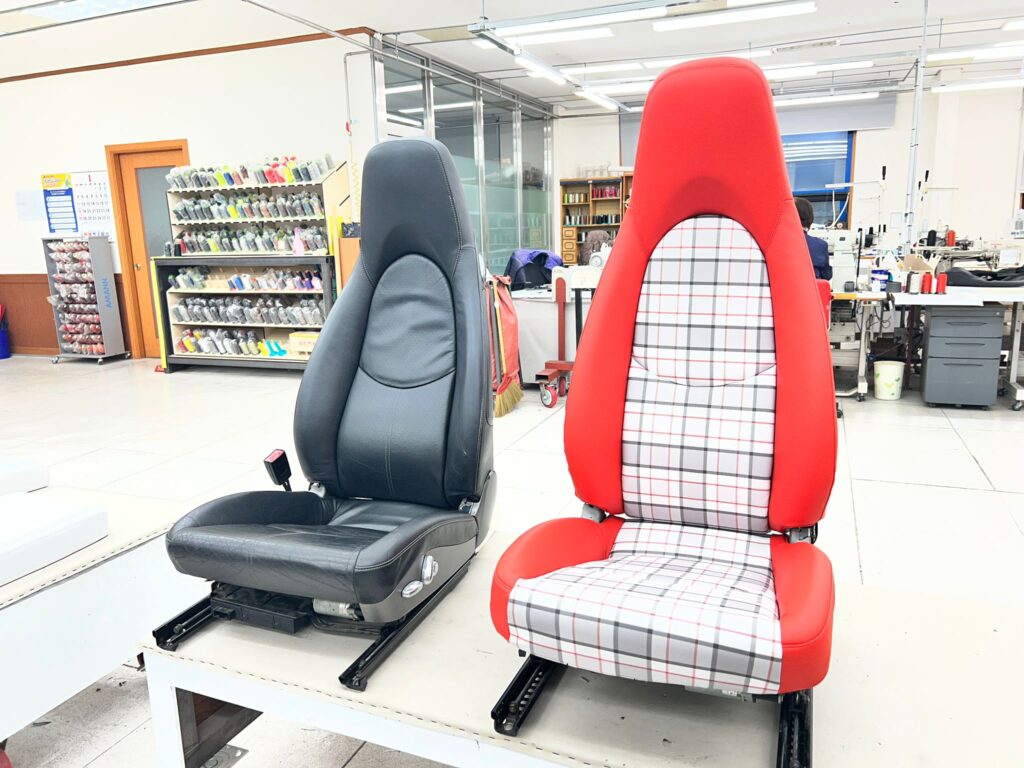 Before and after comparison of a Porsche car seat, the original black leather seat on the left and the reupholstered red leather and plaid seat on the right, displayed side by side in a factory workshop with thread racks and sewing stations in the background.