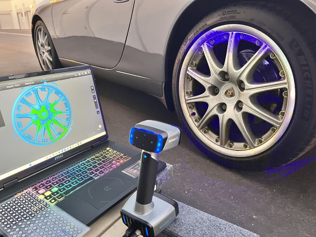 A handheld 3D scanner with blue light projecting onto a Porsche wheel, with a laptop screen showing the rapid scan in progress and the wheel mapped in blue and green point cloud data.