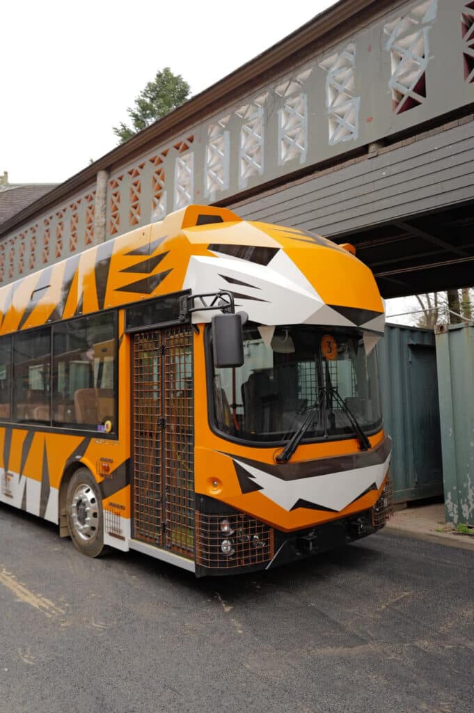 Orange, white, and black tiger striped safari bus with a custom wrapped exterior, parked beneath a covered walkway with wire mesh grille guards on the front.