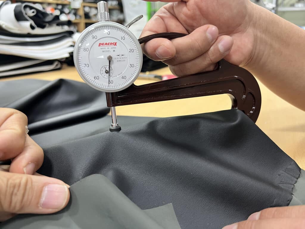 Close up of a dial thickness gauge measuring a piece of gray leather, with hands positioning the fabric and a stack of folded materials in the background.